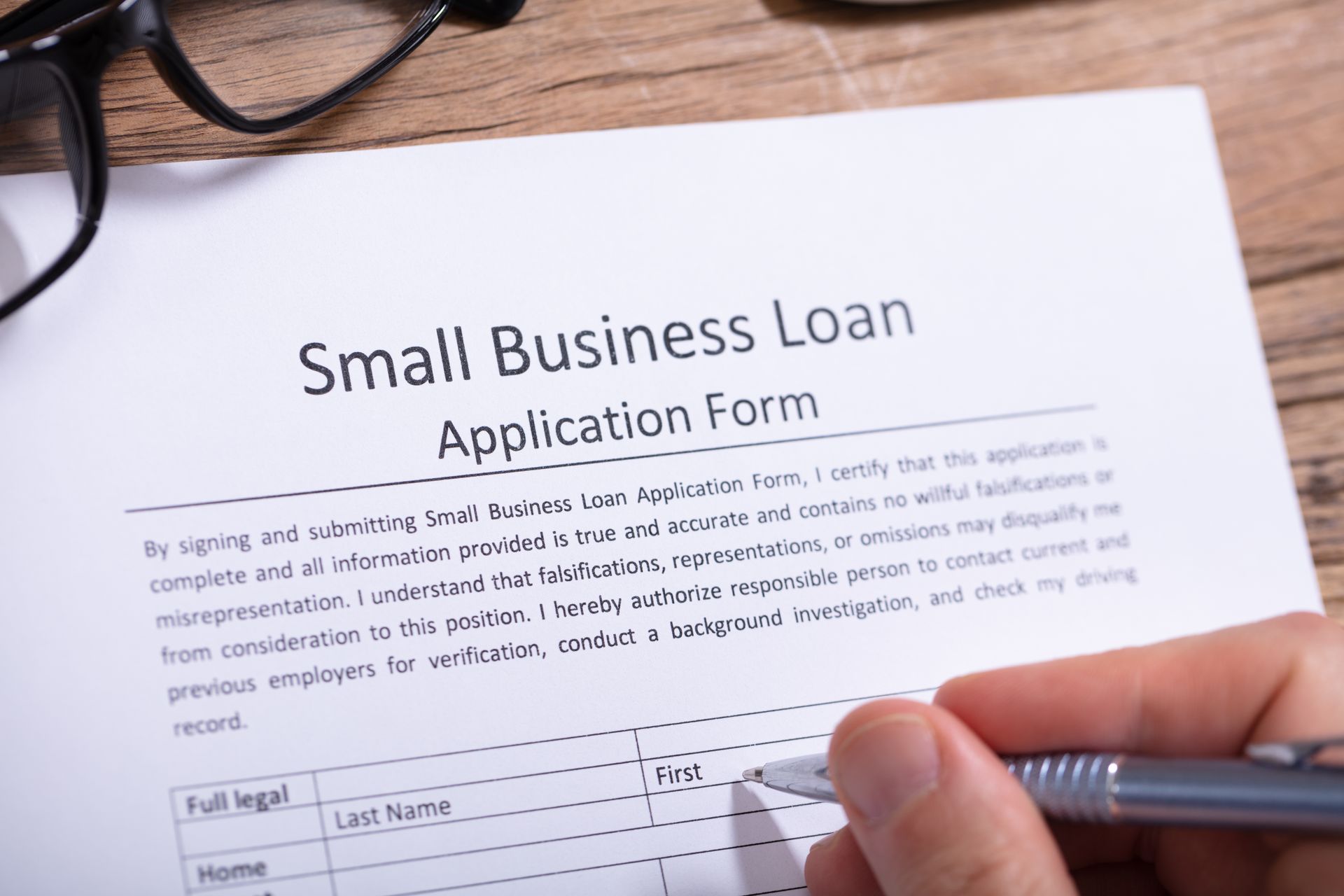 A close-up of a person's hand holding a pen over a Small Business Loan Application form on a wooden desk with glasses.