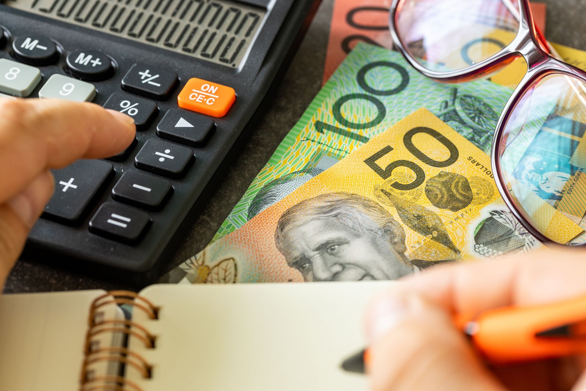 A person using a calculator with Australian cash, glasses, and a notebook on a desk. A person using a calculator with Australian cash, glasses, and a notebook on a desk.