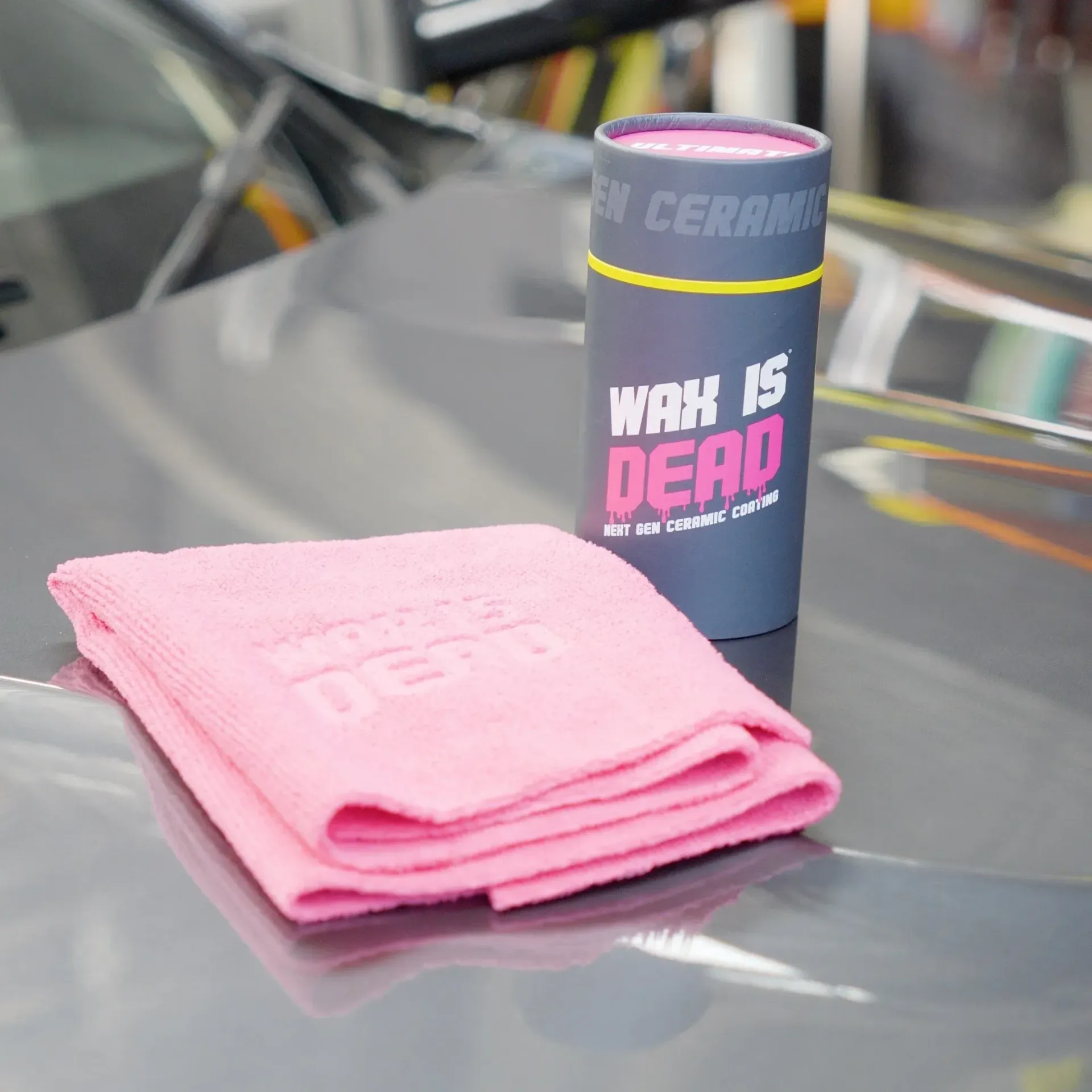 Pink microfiber cloth beside a black “WAX IS DEAD” ceramic container on a car hood