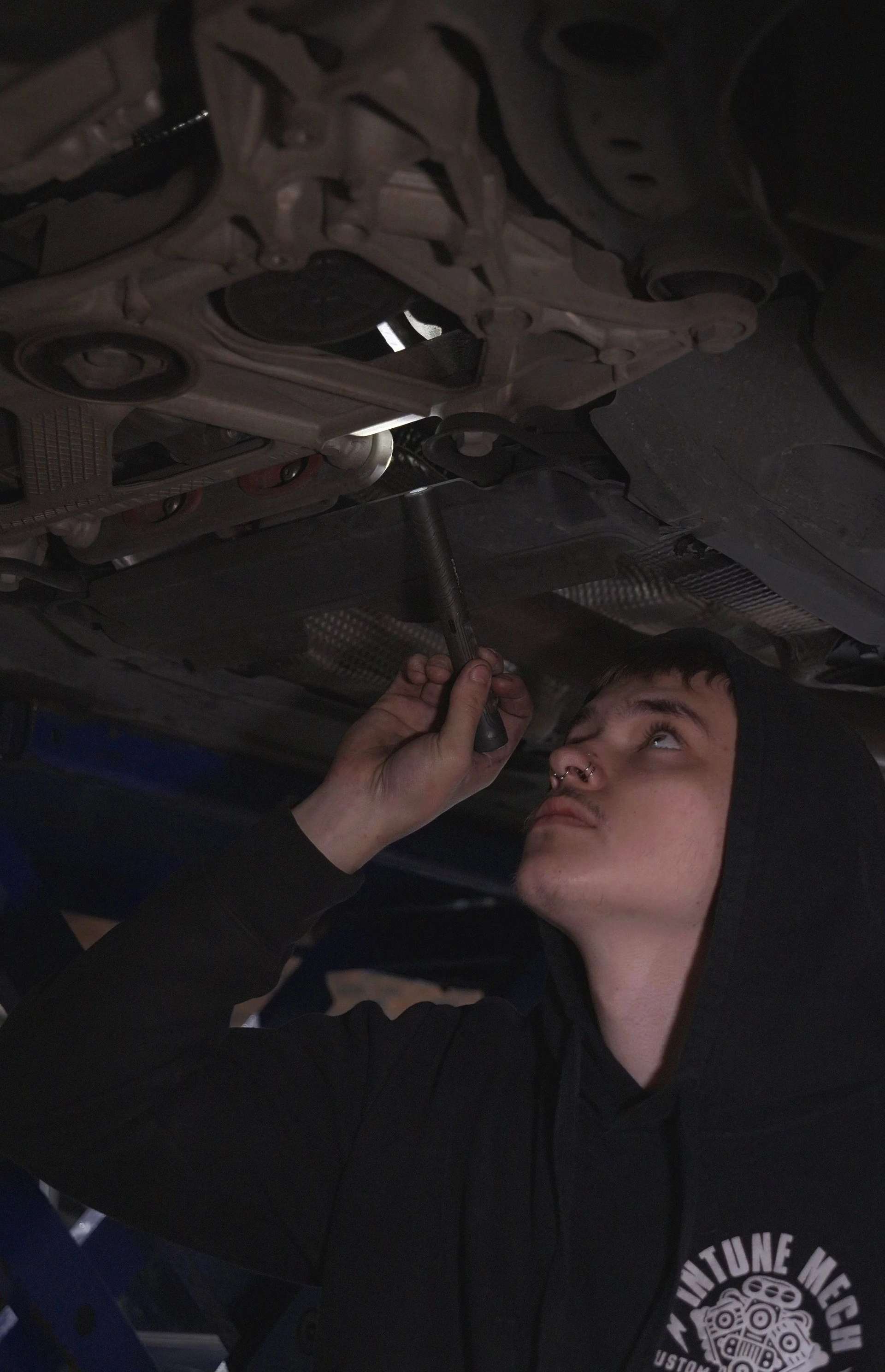 Person working under a vehicle with a flashlight, inspecting the underside in a dim garage