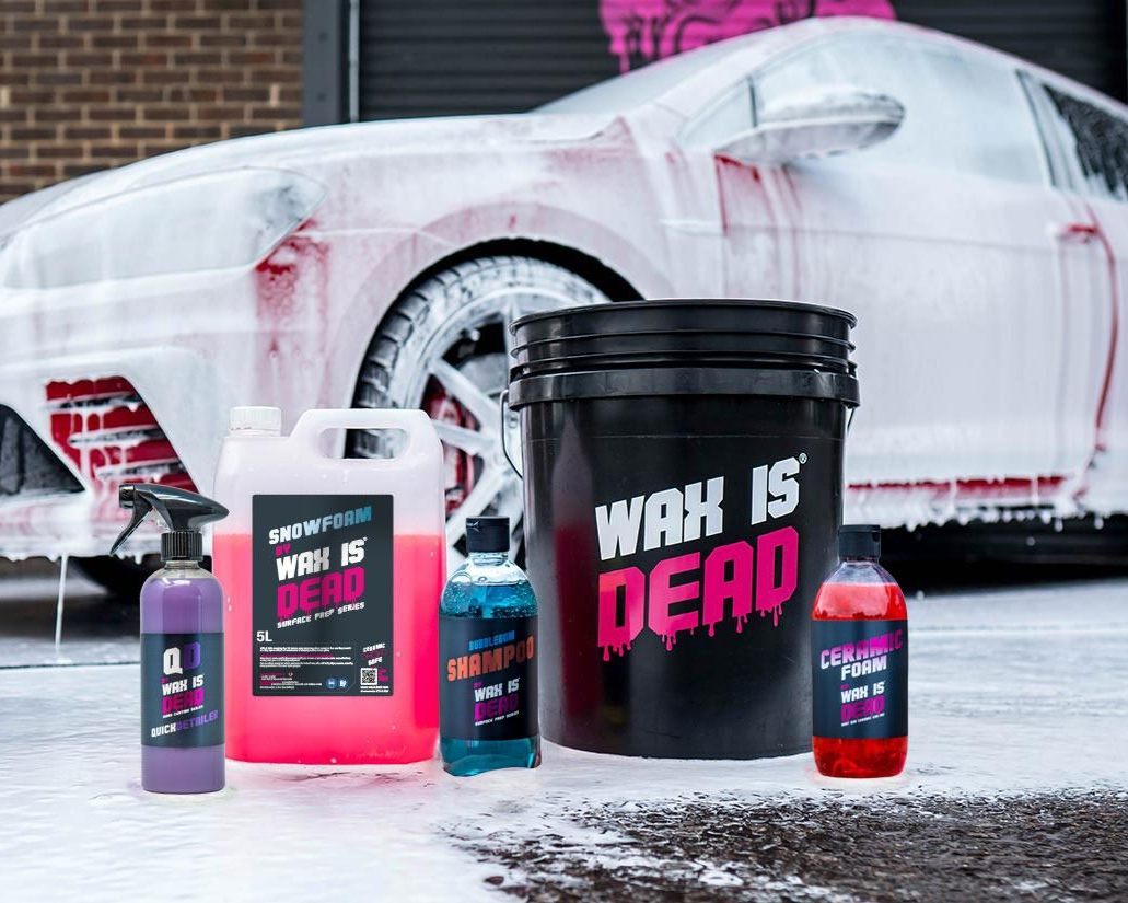 Car wash detailing products in front of a white sports car, with a black bucket labeled “WAX IS DEAD.”