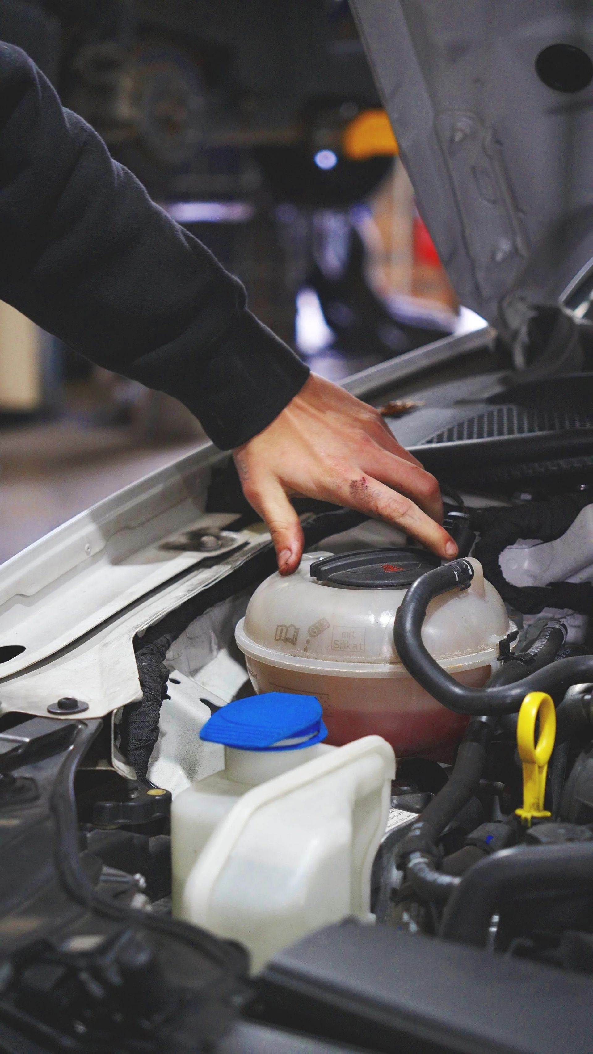 Person inspecting a car engine with hood open, hand near coolant reservoir in an auto shop