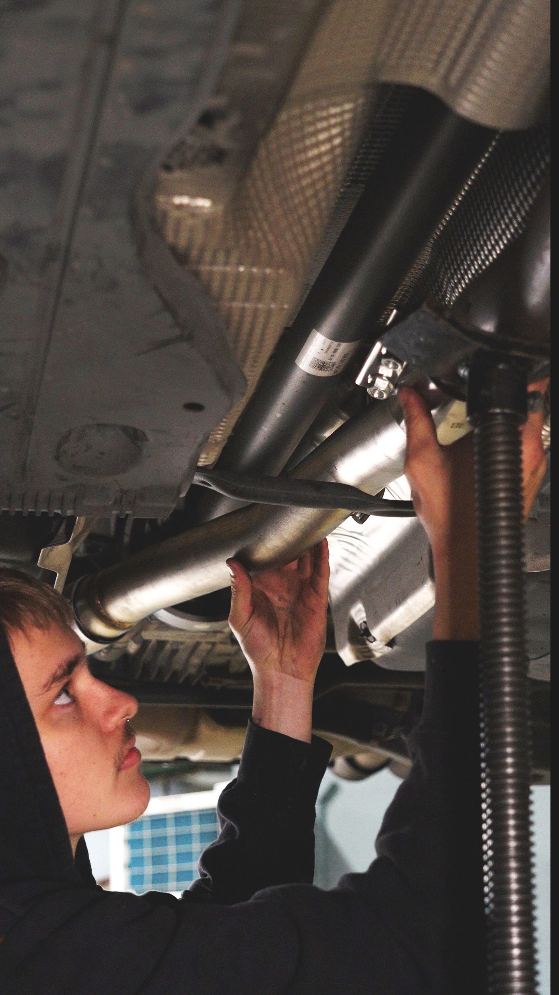 Two people inspect shiny metal pipes and hoses under a vehicle.