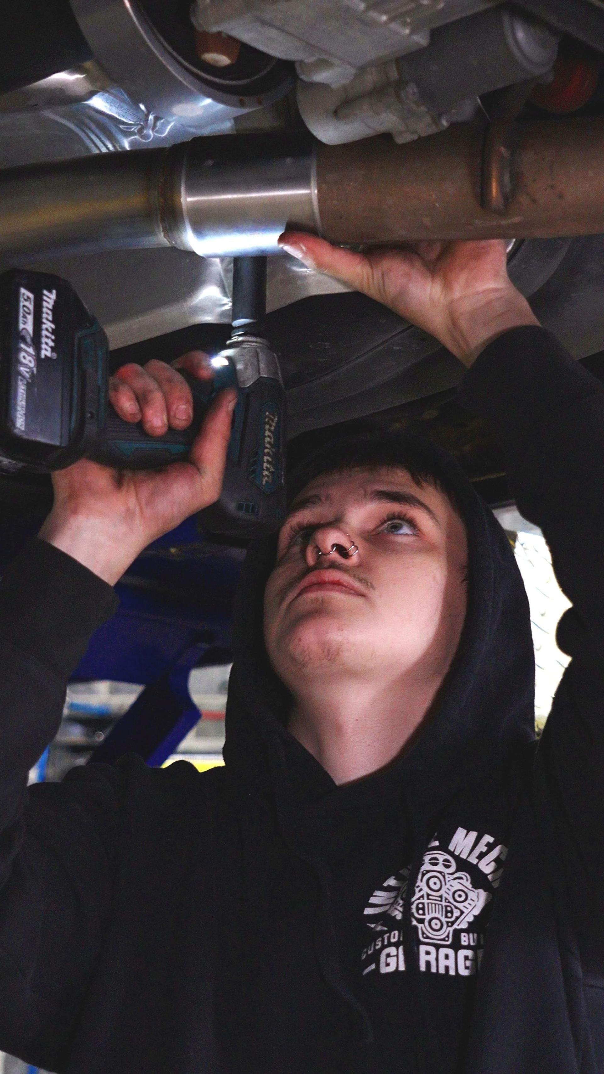 Person using a power tool to work on metal pipes under a vehicle, wearing a black hoodie.