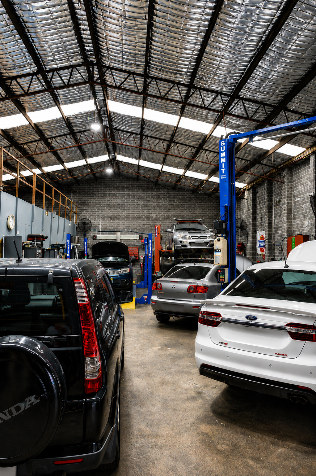 Car repair shop interior with several cars parked under a high metal roof, including a white SUV and black van.