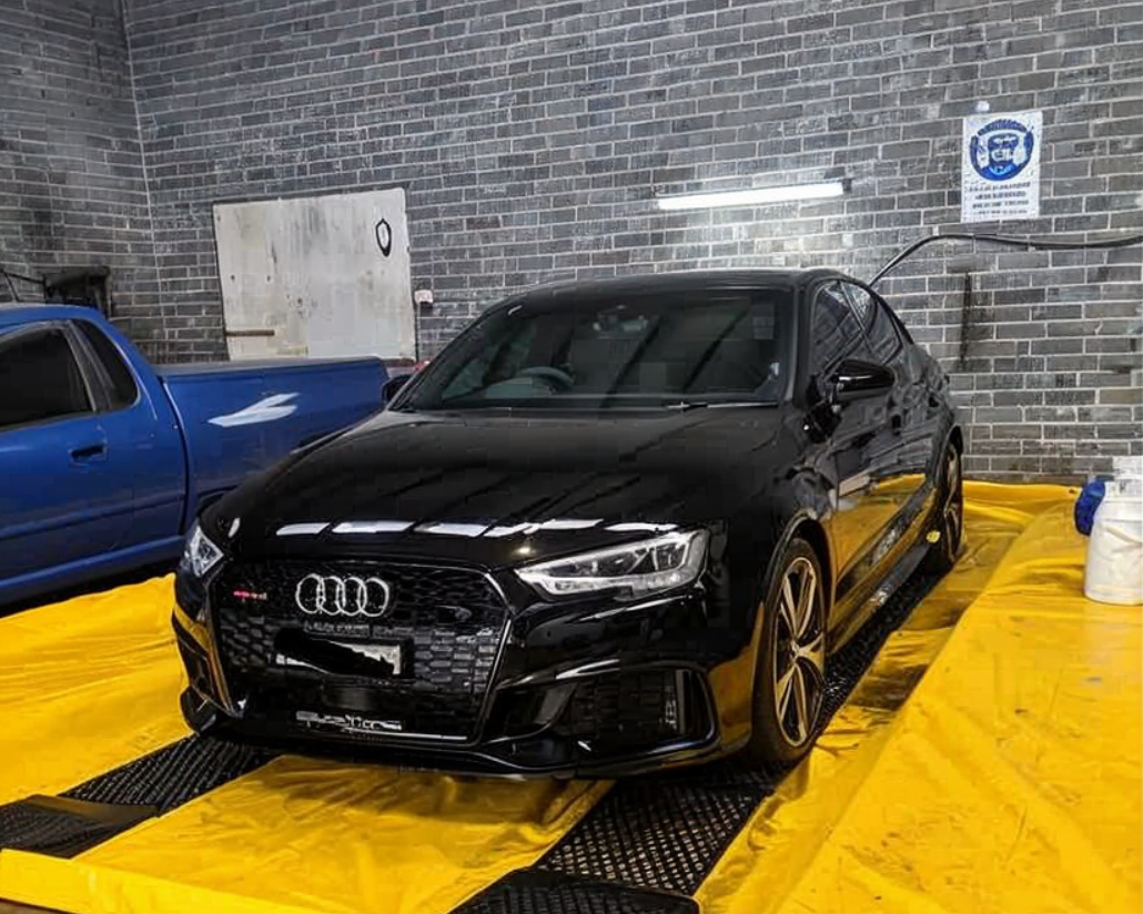 Black Audi sedan parked indoors on a bright yellow floor beside a blue car in a brick-walled garage