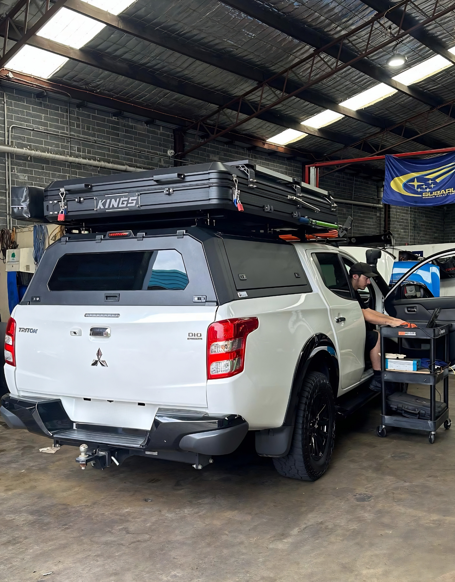White pickup truck in a workshop, with roof cargo and a person working beside it