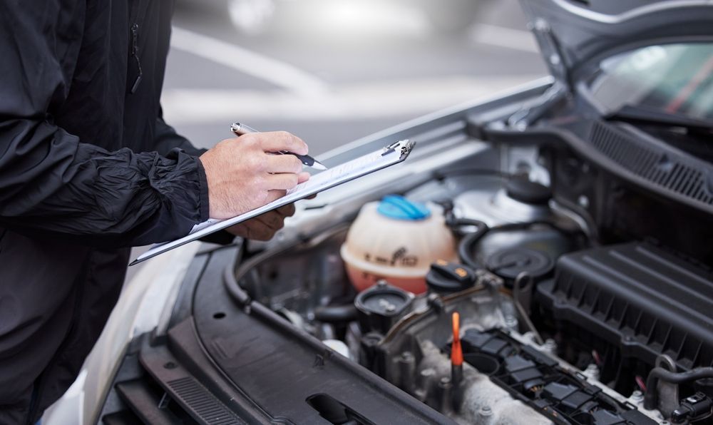 Mechanic writing on a clipboard beside an open car hood with exposed engine components