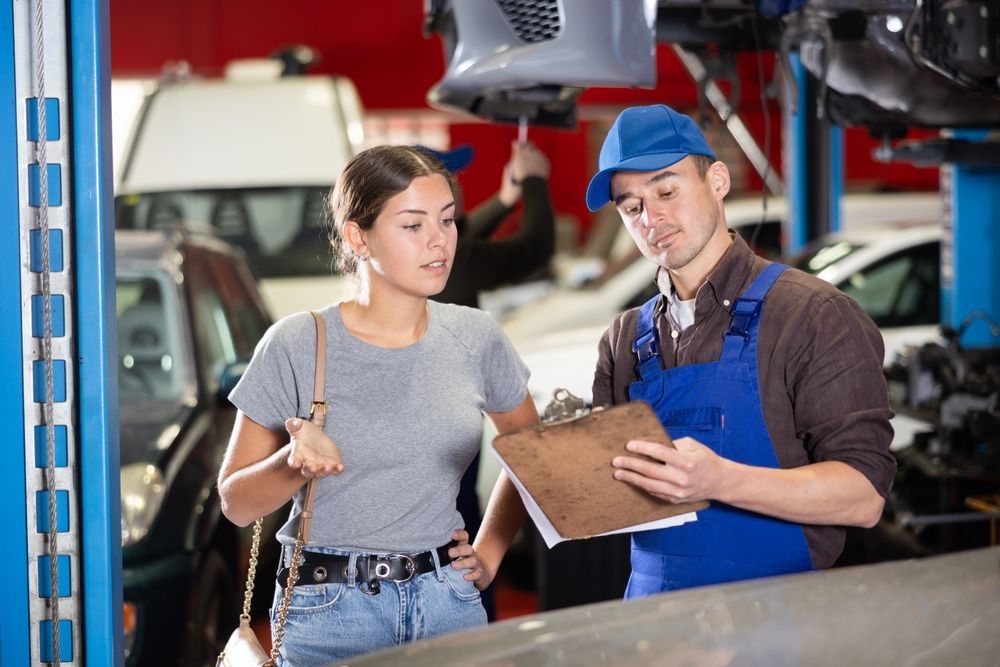 Mechanic in blue coveralls showing a clipboard to a customer in a garage, with cars in the background