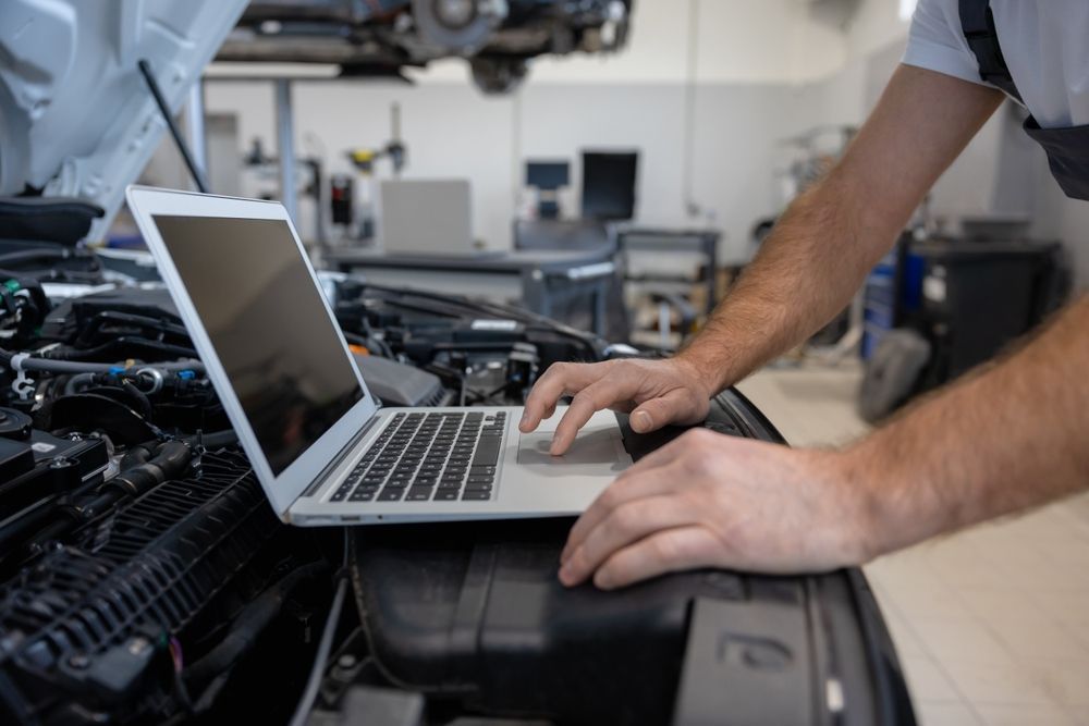 Mechanic using a laptop in a car repair garage, working beside an open engine compartment