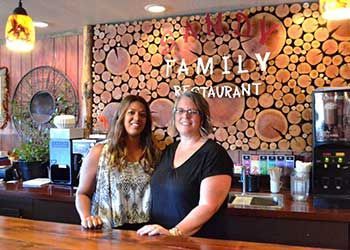 Two women are standing in front of a wooden wall that says family restaurant.