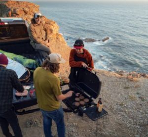 A group of people are standing around a grill on a cliff overlooking the ocean.