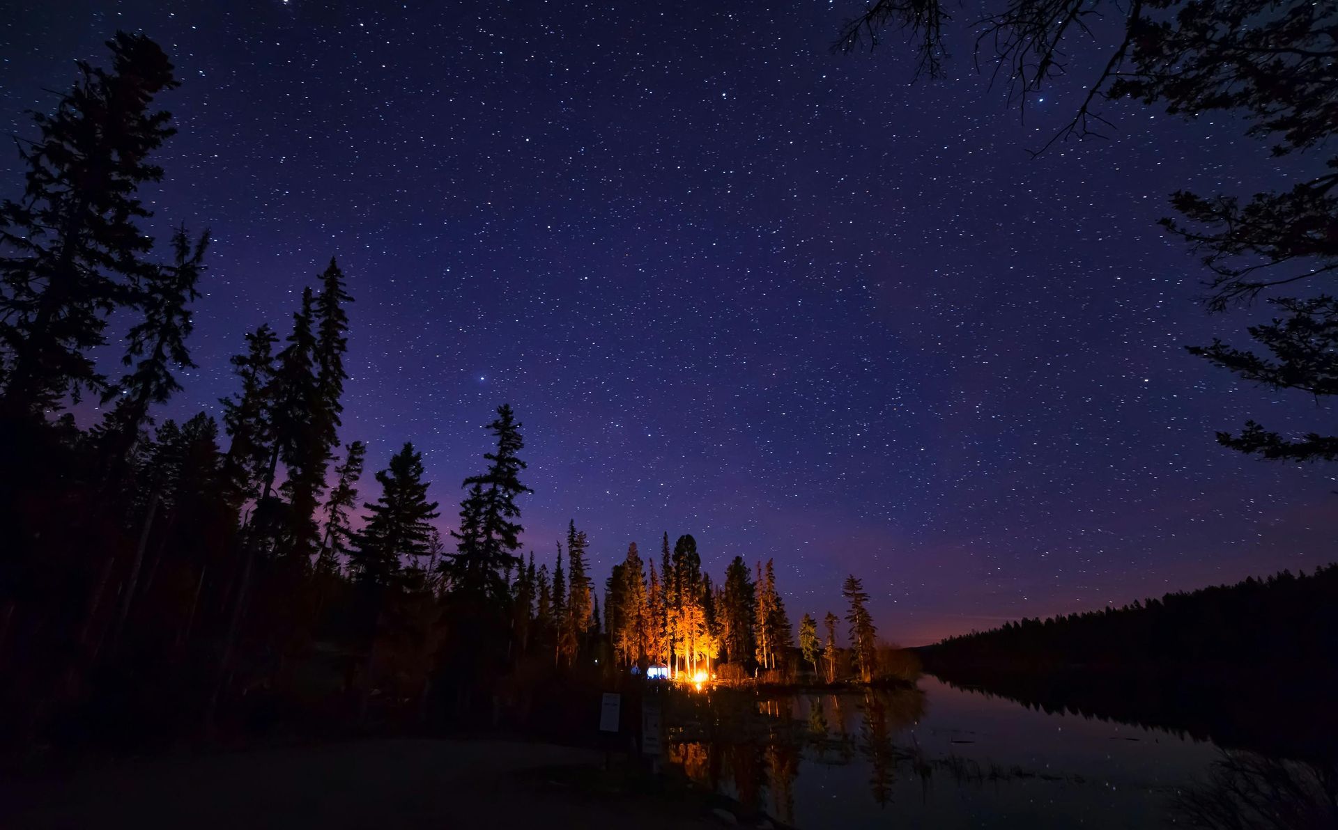 A campfire is lit up in the middle of a lake under a starry night sky.