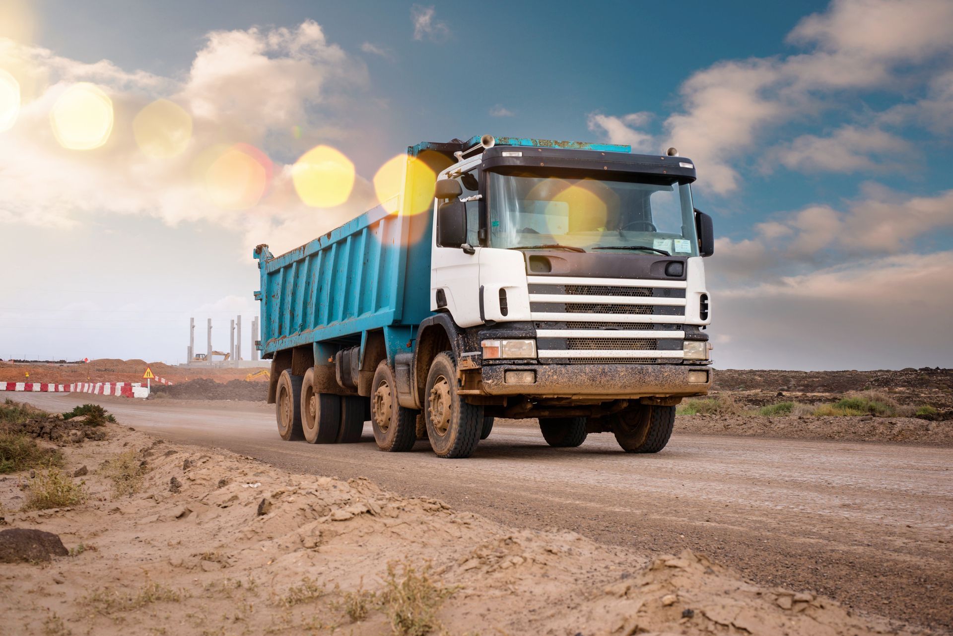 A blue and white dump truck driving on a dirt road under a cloudy sky.