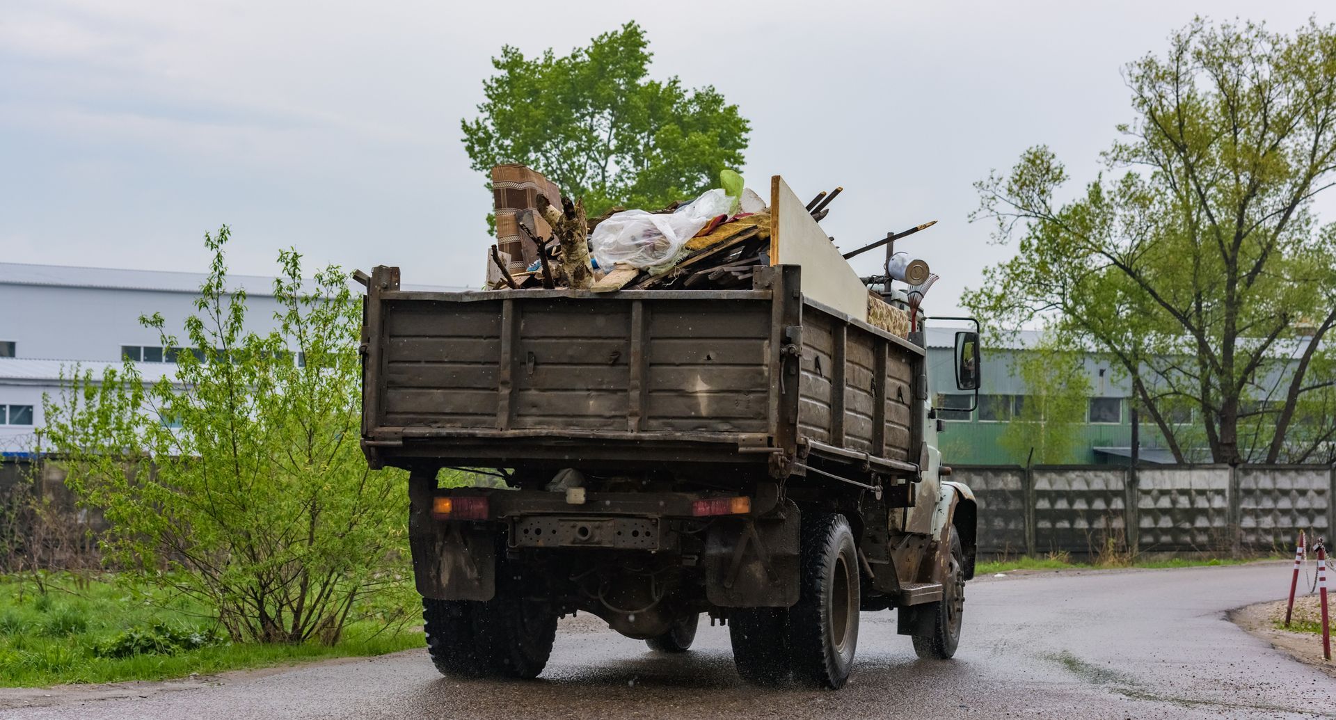 Dark truck filled with debris, driving on a wet road near a building with green trees.