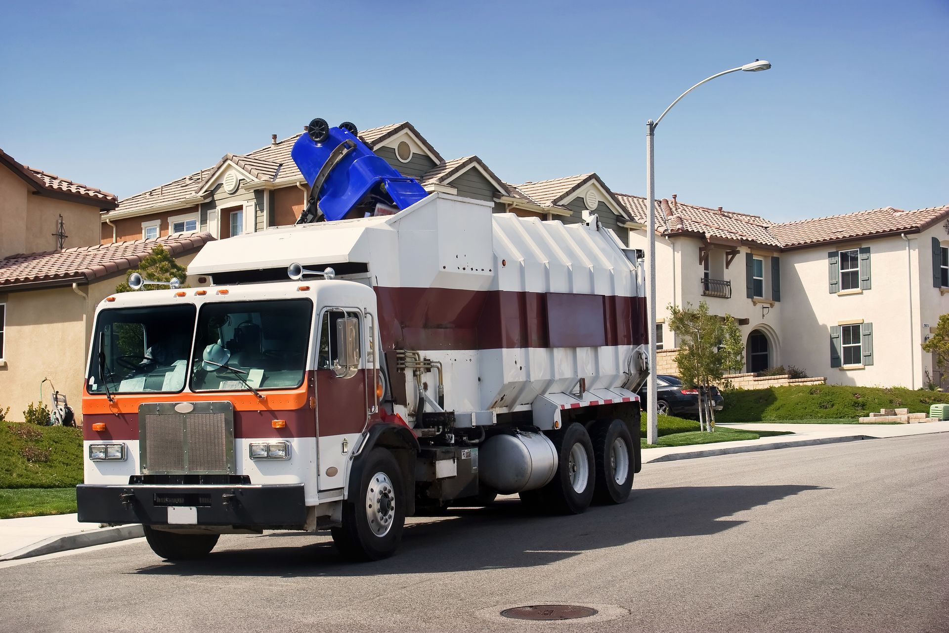 Garbage truck on a residential street, lifting a blue bin. Houses and a street lamp are in the background.