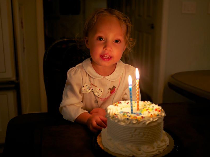 A two-year-old girl smiles with a small cake and two lit candles in front of her
