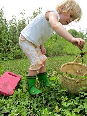 A little girl wearing green rain boots is picking berries in a field.