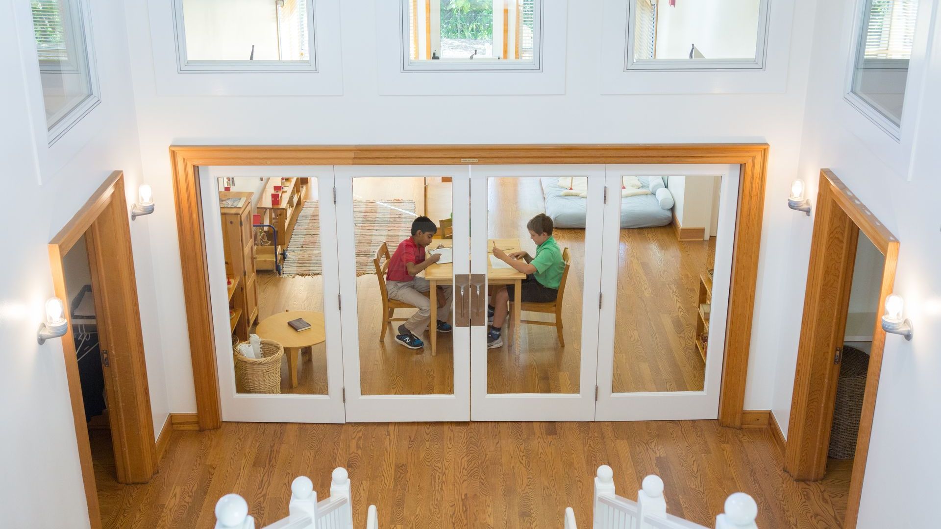 A couple of Montessori children are sitting at a table in a room with a lot of windows.