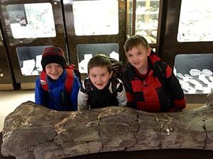 Three young boys are standing next to a large rock.