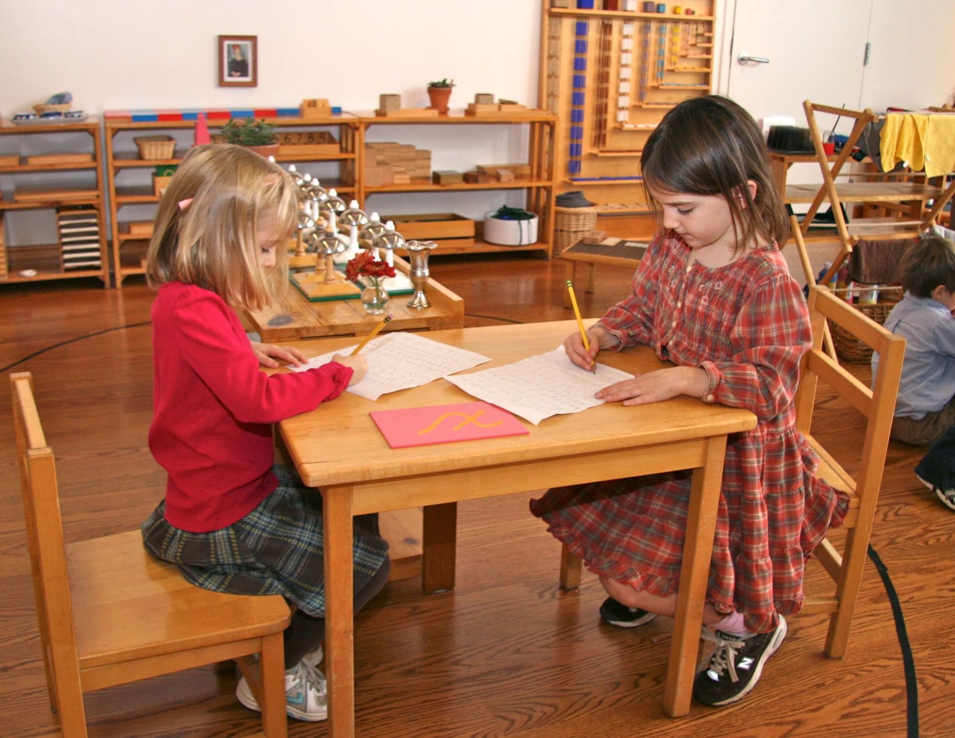 Two young girls in a Montessori Primary Level class work on their cursive at a table together