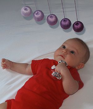 A baby in a red shirt is laying on a bed looking at Montessori mobile