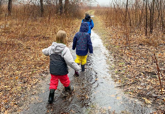 Three children are walking down a muddy path in the woods.