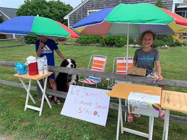 A boy and a girl are selling lemonade under umbrellas in a yard