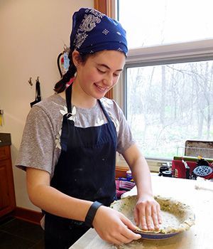 A young girl wearing an apron and a bandana is preparing a pie in a kitchen.