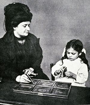 Dr. Montessori closely observes a child working with Metal Insets, one of the many learning materials she designed based on her research.