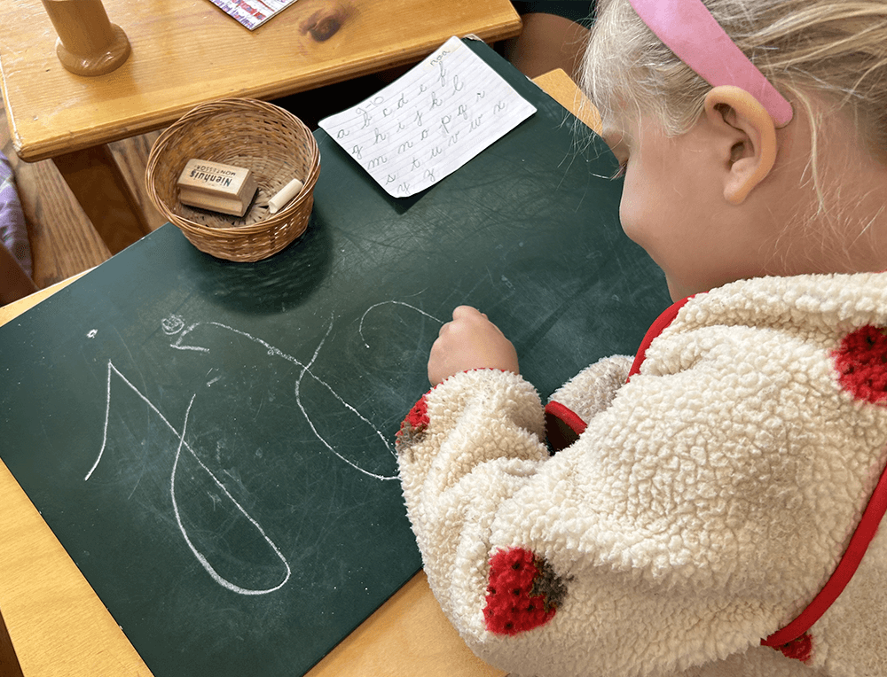 A young Montessori student practices cursive handwriting with a piece of chalk and small blackboard