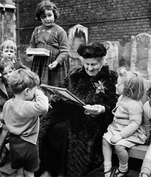 Maria Montessori in old age is reading a book to a group of children.