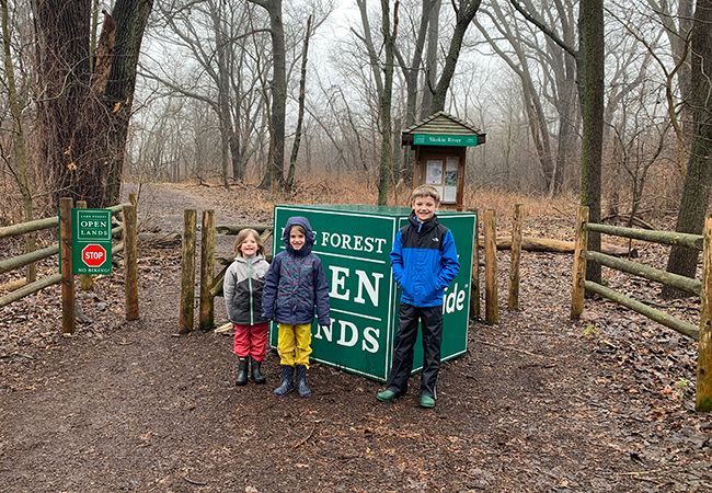 Three children are standing in front of a forest entrance sign.