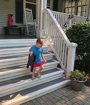 A small child going down the porch stairs of a house with a folding stool under his arm