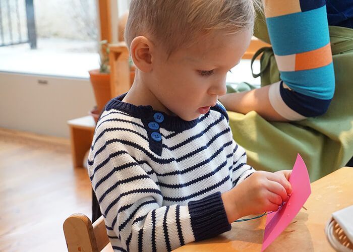 A little boy is sitting at a table sewing with a piece of paper.