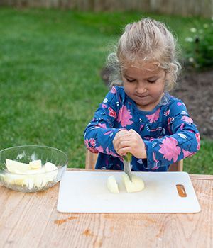 A little girl is cutting an apple on a cutting board.