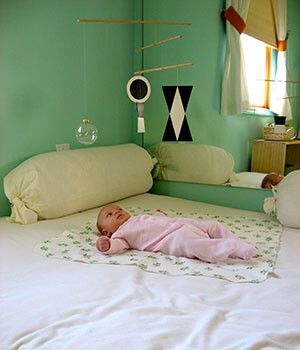 A baby in a pink outfit is laying on a white blanket on a bed looking at a Montessori mobile.