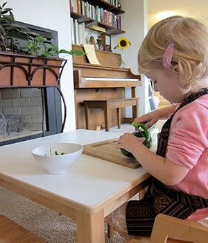 A little girl is sitting at a table cutting a cucumber.