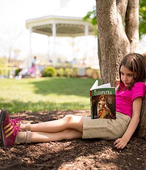 A girl reading a book sitting down a tree.