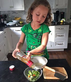 A little girl is pouring dressing into a bowl of vegetables.