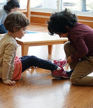 A boy is helping a little girl tie her shoes on the floor.