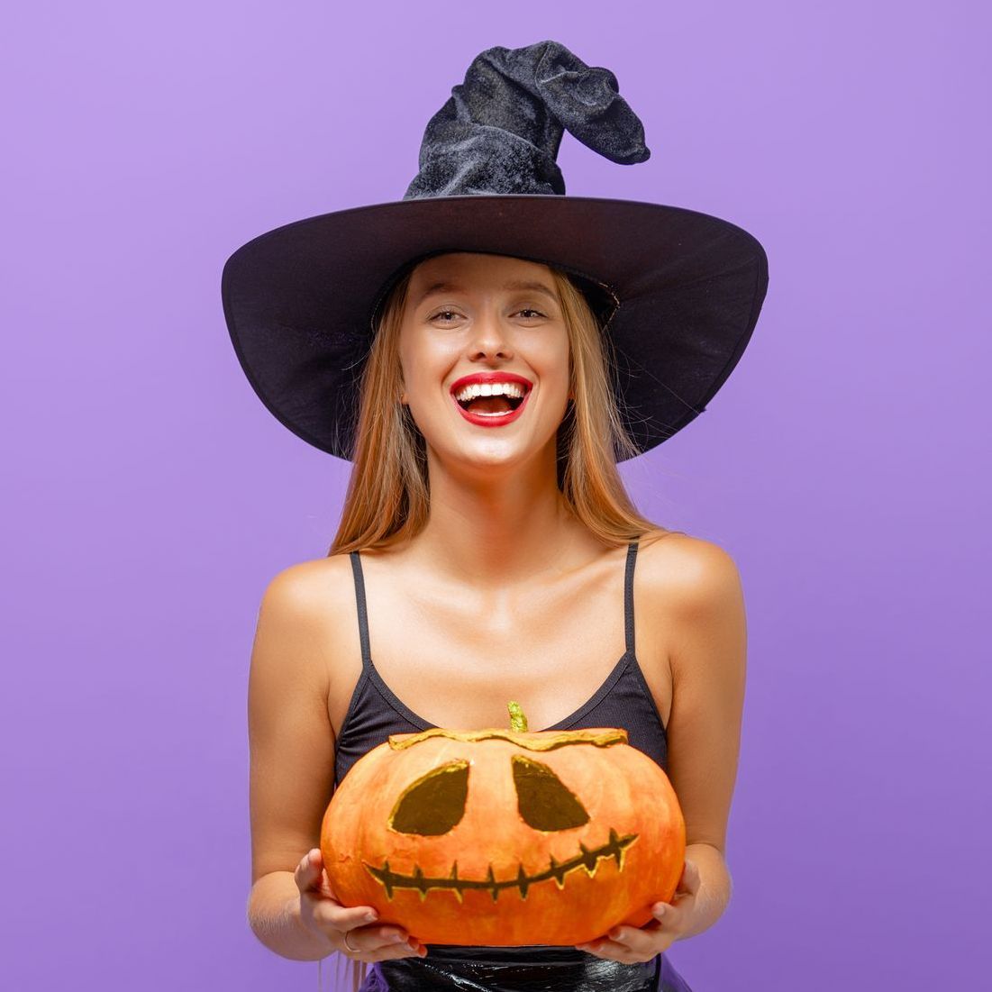 A woman in a witch hat is holding a pumpkin with a face carved into it.