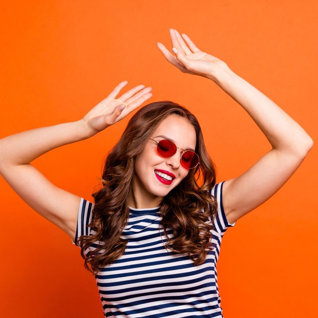A woman wearing sunglasses and a striped shirt is dancing with her arms in the air.