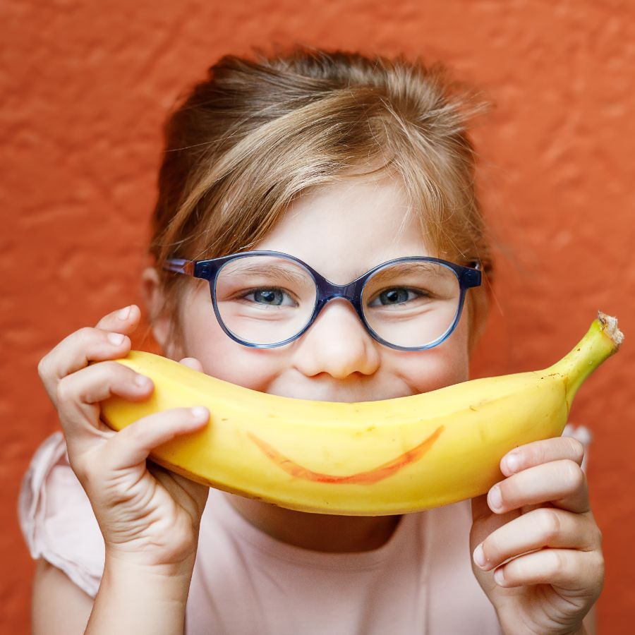 A little girl wearing glasses is holding a banana with a smiling face painted on it.