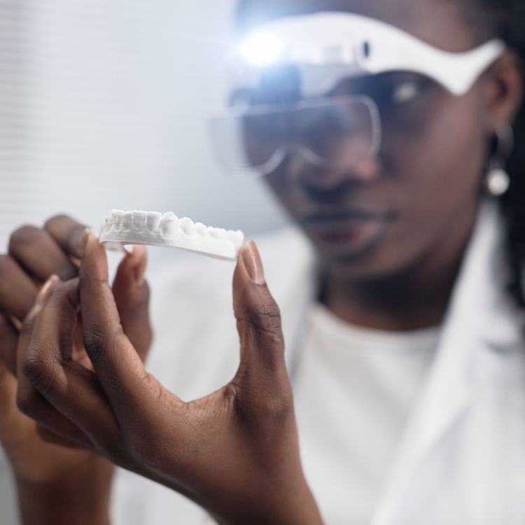 A woman in a lab coat and goggles is holding a model of teeth.