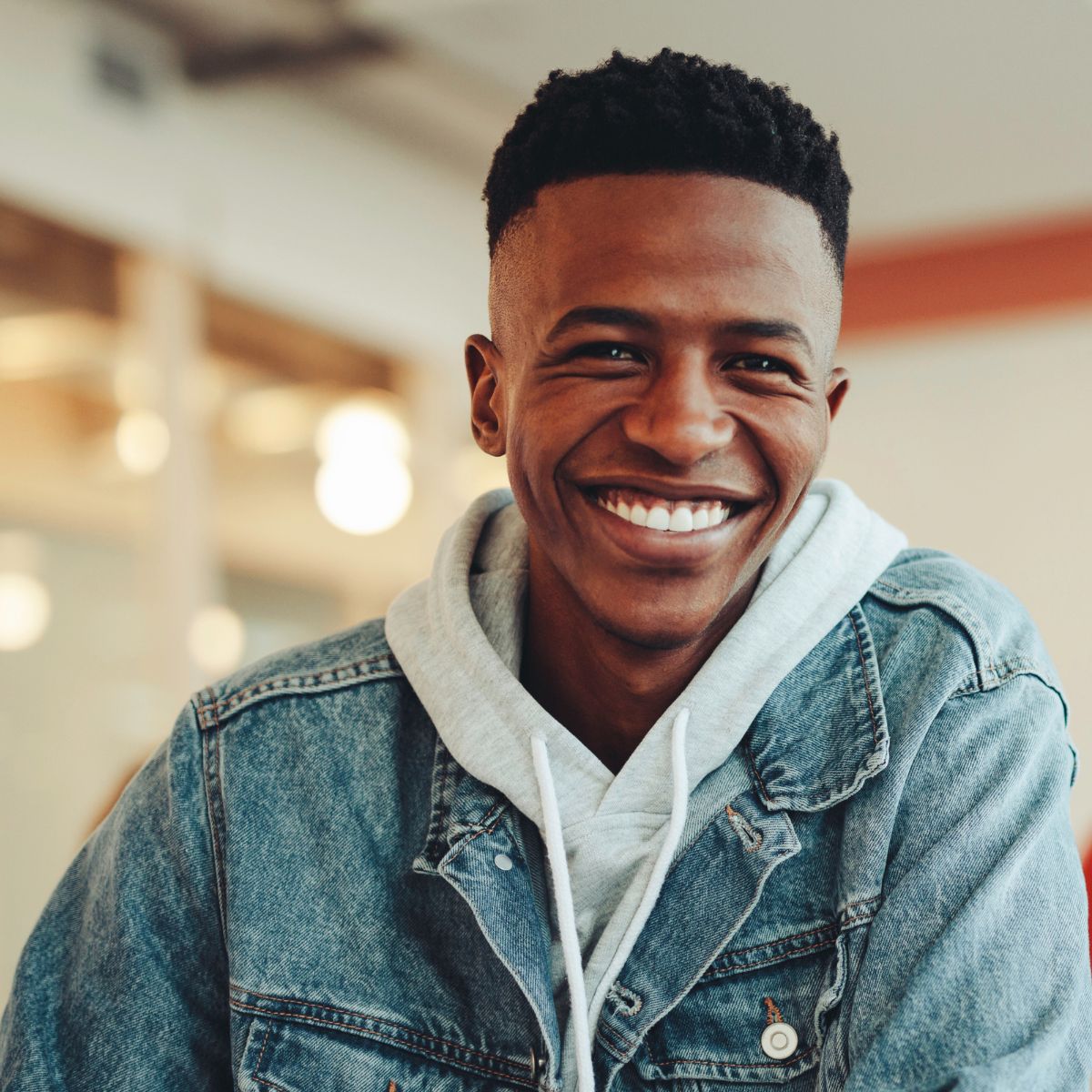 A young man wearing a denim jacket and a grey hoodie is smiling