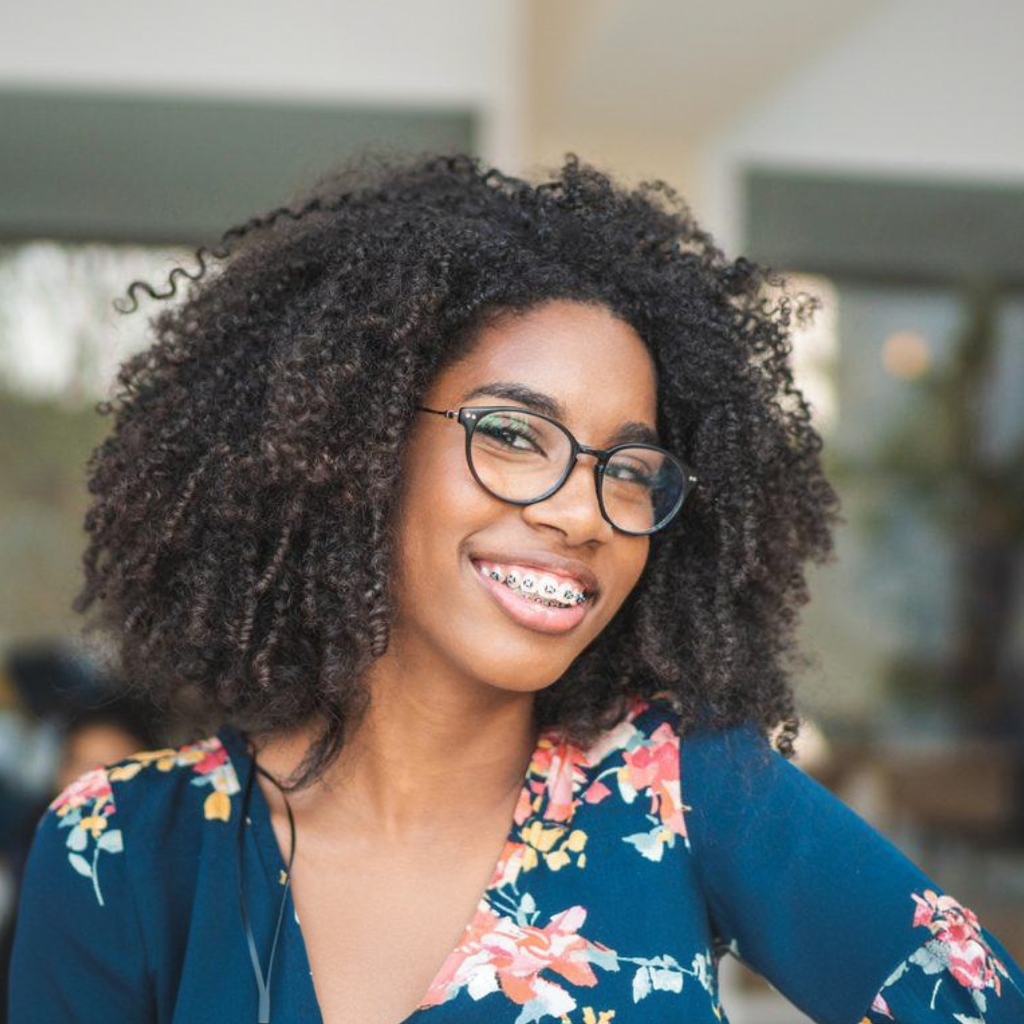 Woman with glasses and braces smiles, wearing a floral top with curly hair.