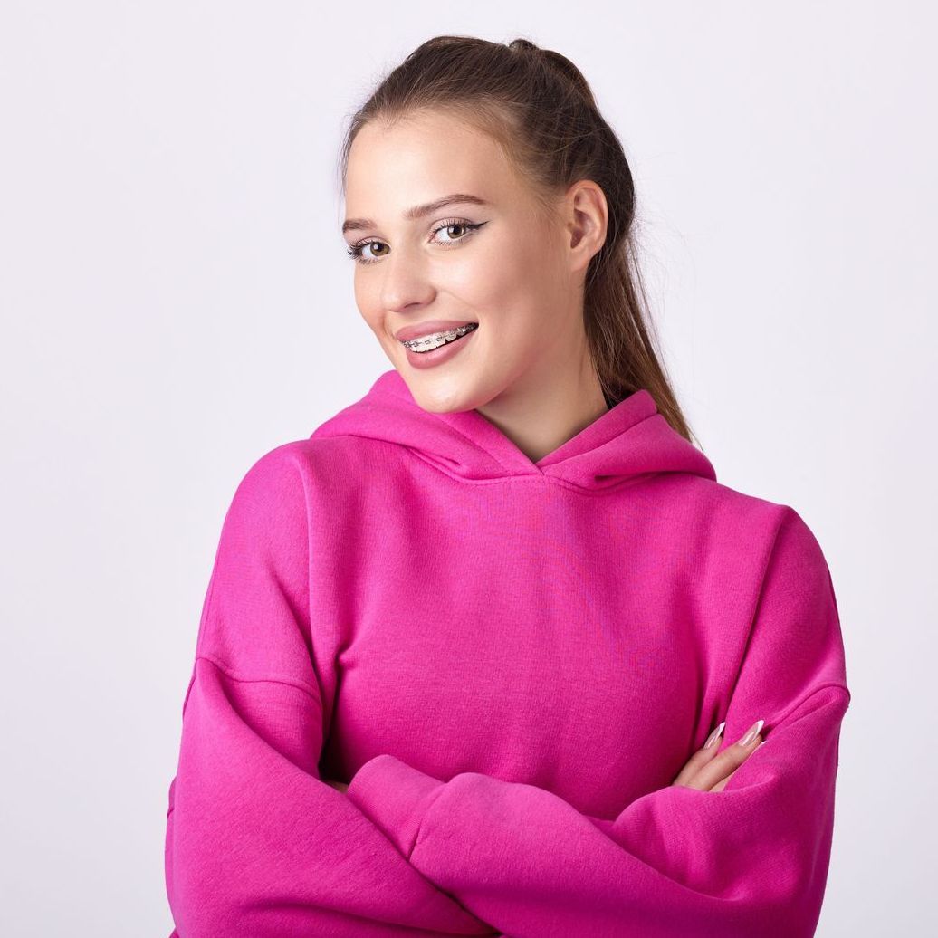 A woman wearing braces and a pink hoodie is standing with her arms crossed.