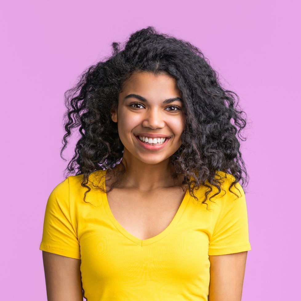 A woman with curly hair is wearing a yellow shirt and smiling.