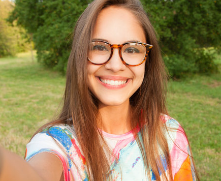 Woman with glasses smiling, taking a selfie outdoors with green foliage in the background.